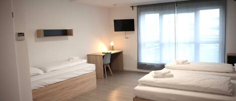A modern hotel room with two single beds, a desk, a chair, a TV mounted on the wall, and a large window with curtains.