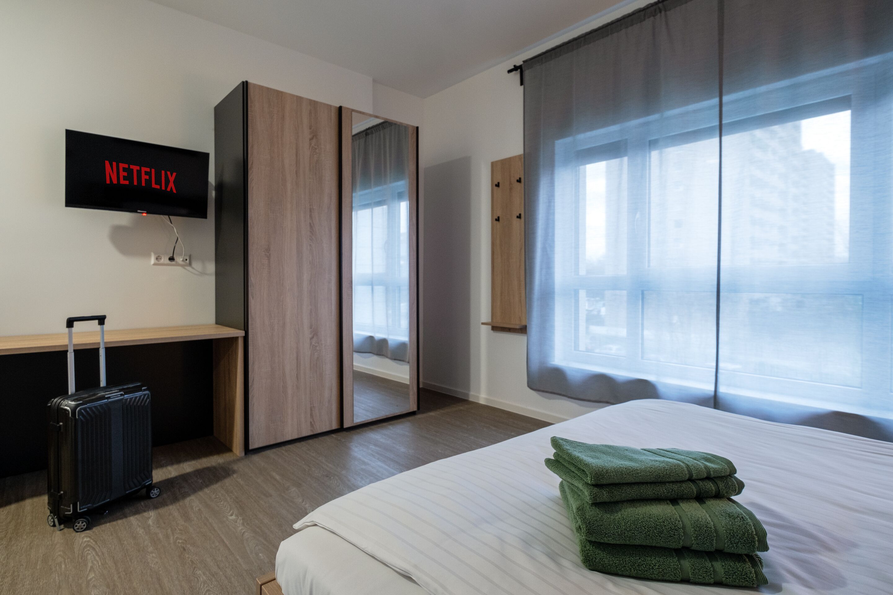 Habitación de hotel con cama, escritorio, maleta, televisor mostrando 'NETFLIX' y un ventanal con cortinas.