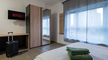 Habitación de hotel con cama, escritorio, maleta, televisor mostrando 'NETFLIX' y un ventanal con cortinas.
