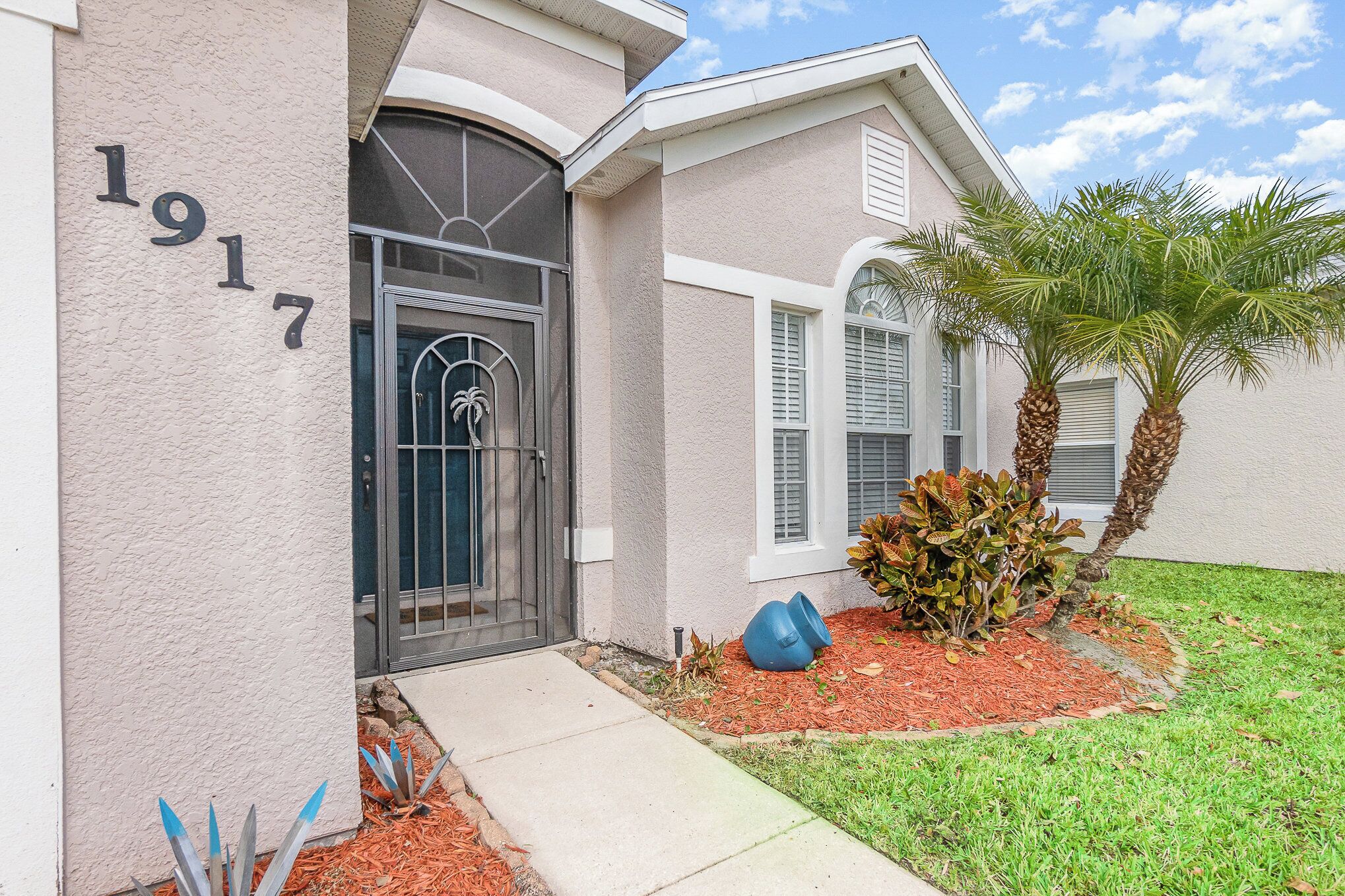 A house with a number '1917' on the wall, a front door, and a small garden with a palm tree and a plant.