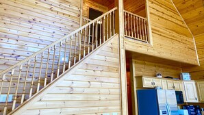 A wooden cabin interior with a staircase leading to a lofted area, a kitchen with modern appliances, and wooden walls and ceilings.