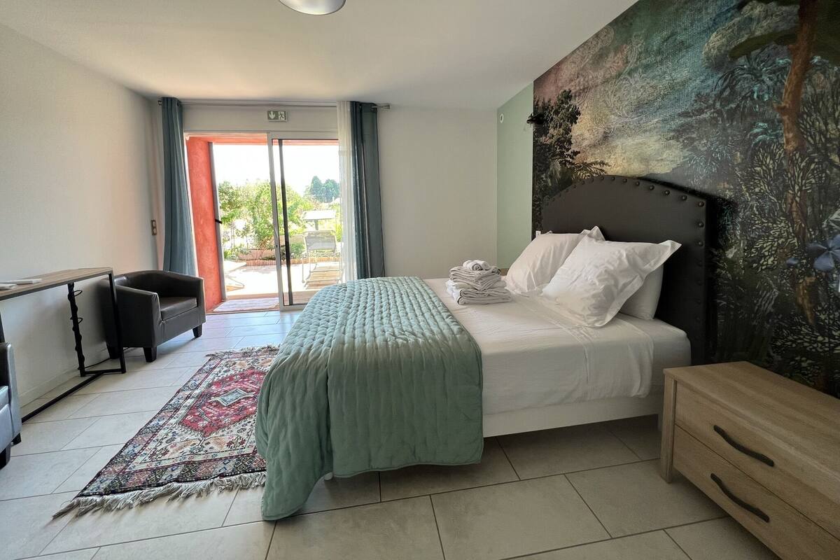 Un dormitorio con cama, mesita de noche, escritorio, silla, un ventanal grande y un cuadro en la pared.