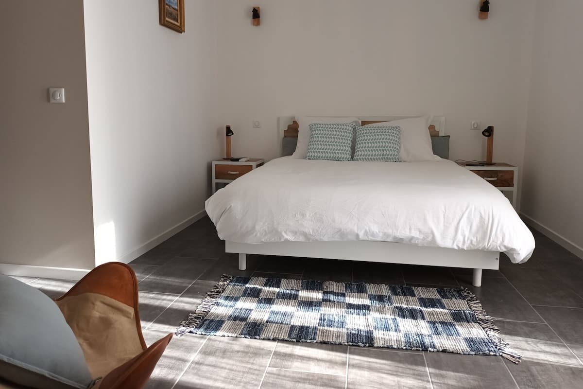 Un dormitorio con una cama grande, dos mesitas de noche, una lámpara auxiliar y una alfombra estampada, además de un cuadro en la pared.