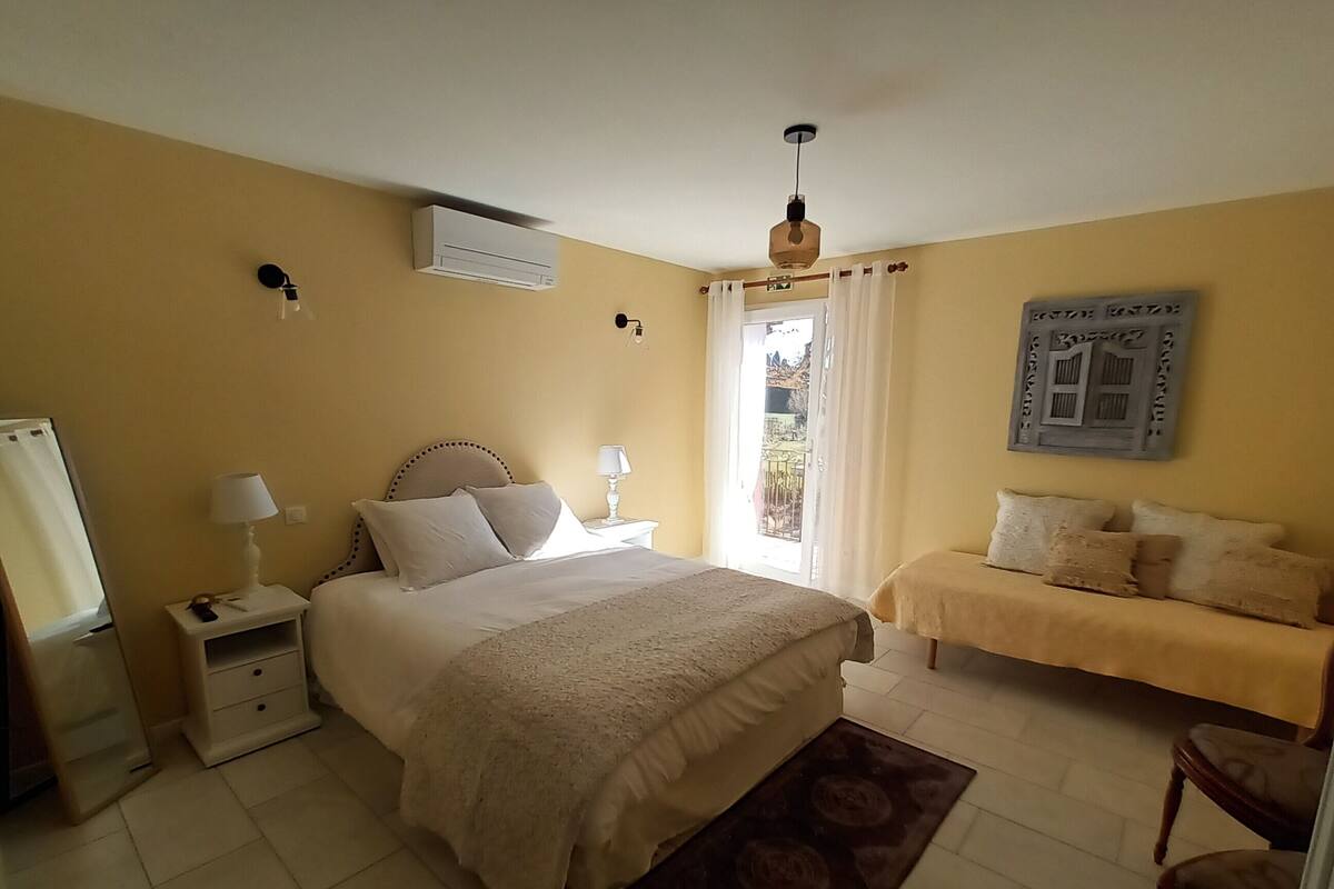 Un dormitorio con una cama grande, un sofá más pequeño, una mesita de noche, una lámpara, un equipo de aire acondicionado y una ventana con cortinas.