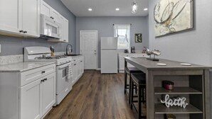 A modern kitchen with white cabinetry, a gas stove, and a refrigerator. There is a wooden dining table with bar stools and a decorative shelf with the word "beth" on it.