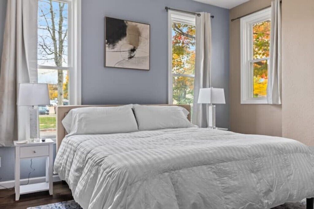 Quarto com cama, duas mesas de cabeceira, uma obra de arte na parede e janelas com cortinas.