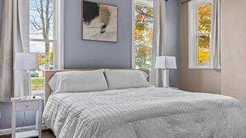 Quarto com cama, duas mesas de cabeceira, uma obra de arte na parede e janelas com cortinas.