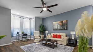 A modern living room with a ceiling fan, a sofa with red cushions, a coffee table, and a large window with curtains.