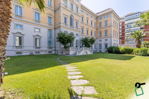 A courtyard with a stone pathway leading to a bridge, surrounded by a well-maintained lawn and buildings with classical architecture.
