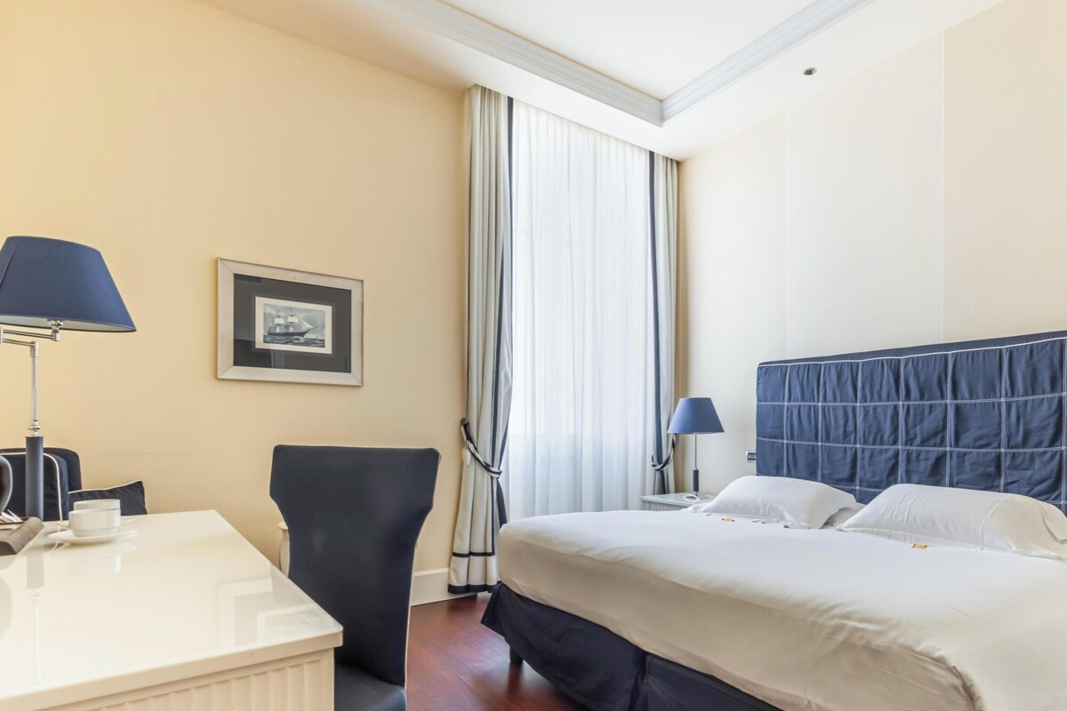 Quarto de hotel com cama, abajures, escrivaninha com luminária, cadeira, quadro na parede e janela com cortinas.