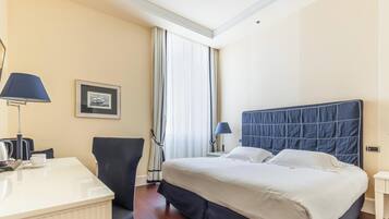 Een hotelkamer met een bed, bedlampjes, een bureau met een lamp, een stoel, een lijst met een foto aan de muur en een raam met gordijnen.