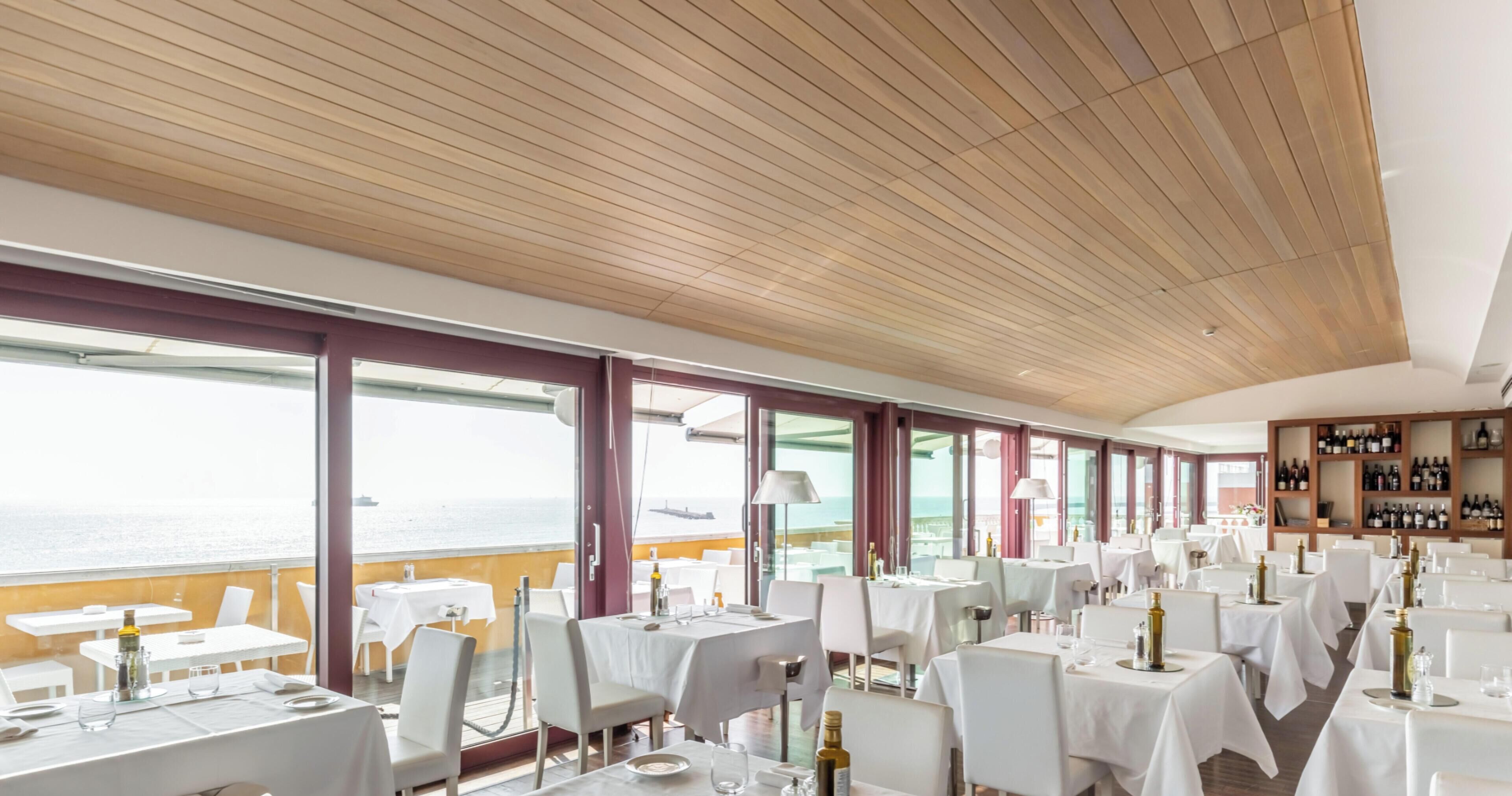 A restaurant with tables set for dining, large windows offering a view of the sea, and a wooden ceiling.