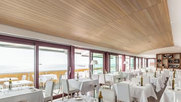 Un restaurant avec des tables dressées pour le service, de grandes fenêtres donnant sur la mer et un plafond en bois.