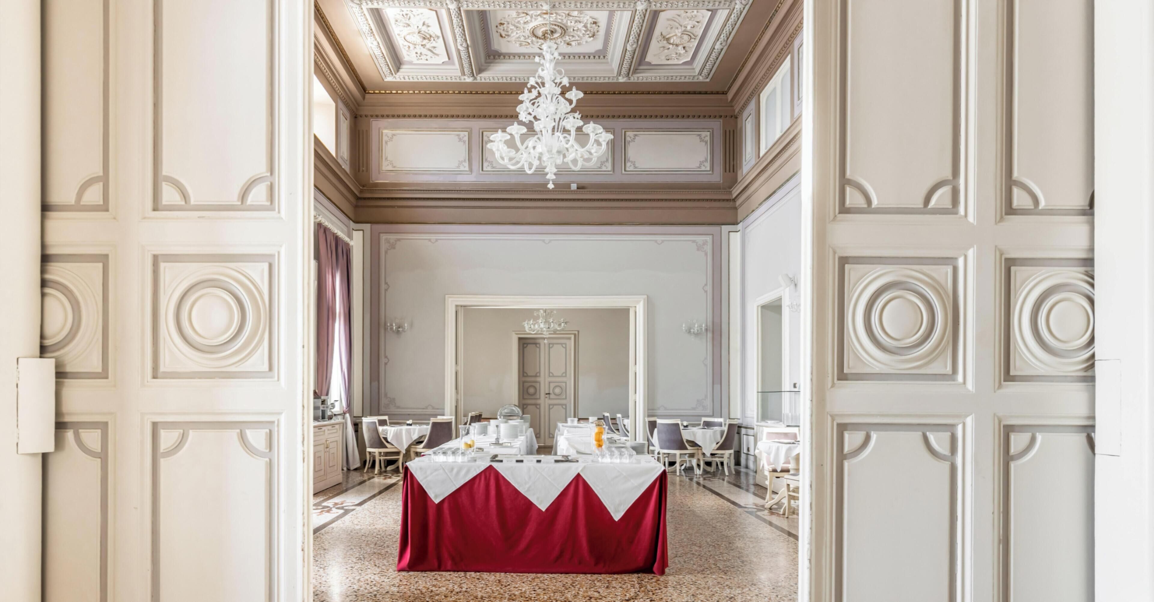 A formal dining room with a chandelier, a red tablecloth, and a classic interior design.