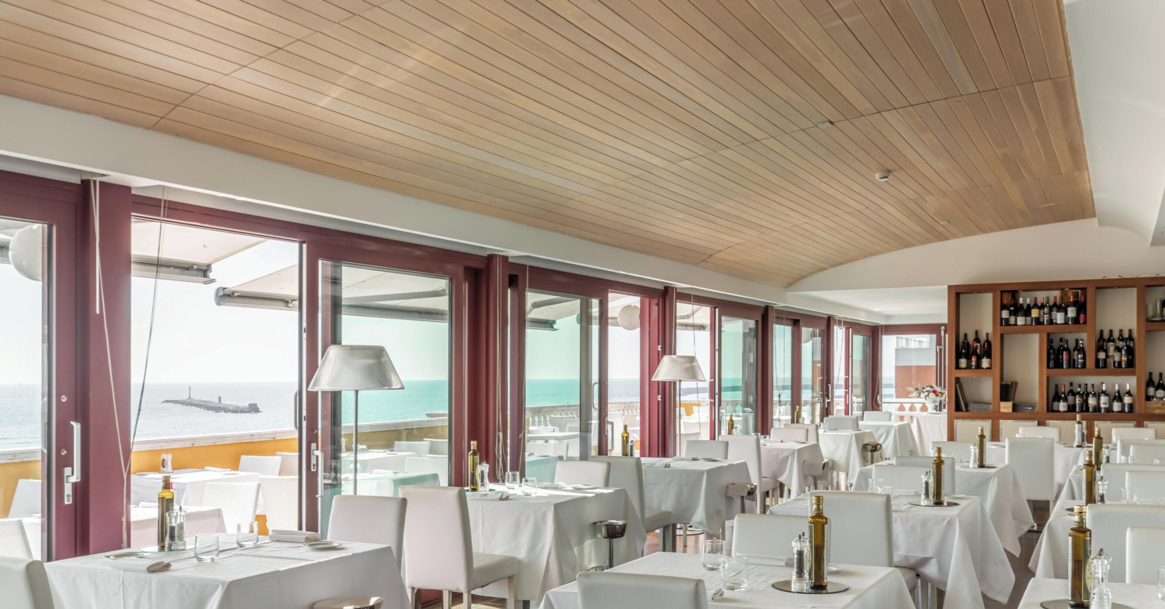 Un restaurant avec des tables et des chaises blanches, de grandes fenêtres donnant sur la mer et un plafond en bois.