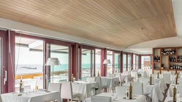 Un restaurant avec des tables et des chaises blanches, de grandes fenêtres donnant sur la mer et un plafond en bois.