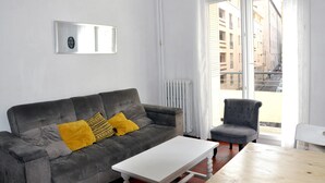 Un salon moderne avec un canapé gris, une table basse blanche et un fauteuil gris. Une grande fenêtre offre une vue sur les bâtiments à l’extérieur.
