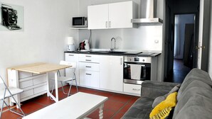 Une cuisine compacte avec des meubles blancs, un four à micro-ondes, une machine à café et un four. On y trouve une table à manger avec des chaises et un canapé dans l’espace salon.