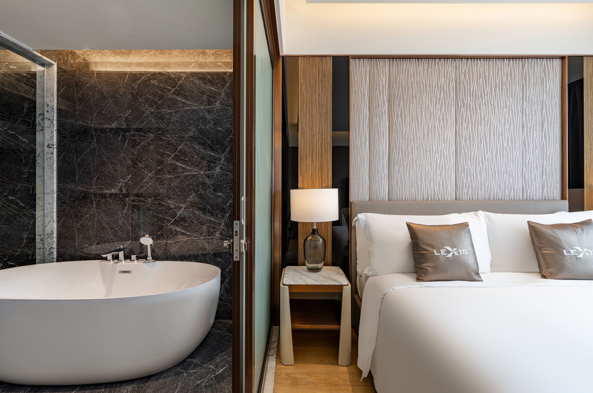 Camera d'albergo moderna con una grande vasca da bagno ovale, un comodino con una lampada e un letto con due cuscini.