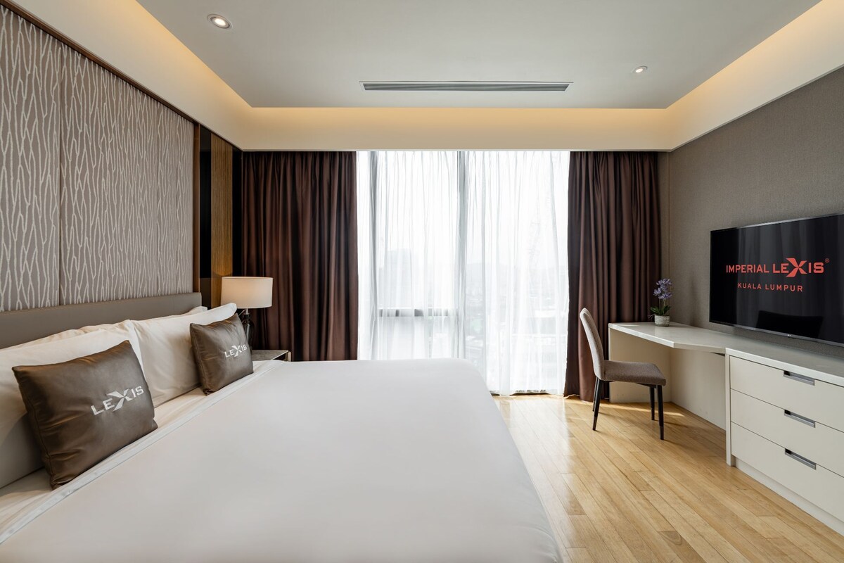 A hotel room with a large bed, a desk with a TV, and a view of the city through the window.