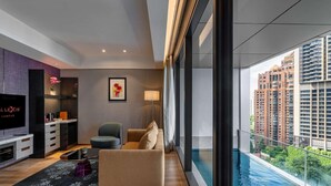 A modern living room with a flat-screen TV, a sofa, a chair, a dining area, and a large window overlooking a cityscape and a swimming pool. - Imperial Lexis Kuala Lumpur (Kuala Lumpur)