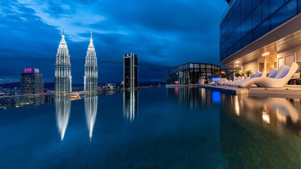 Una piscina infinita en la azotea con vistas a las Torres Petronas, un edificio moderno con fachada de vidrio, y un área de descanso con sillas blancas.