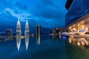 Een dakzwembad met oneindige horizon en uitzicht op de Petronas Towers, een modern gebouw met een glazen gevel, en een lounge met witte stoelen.