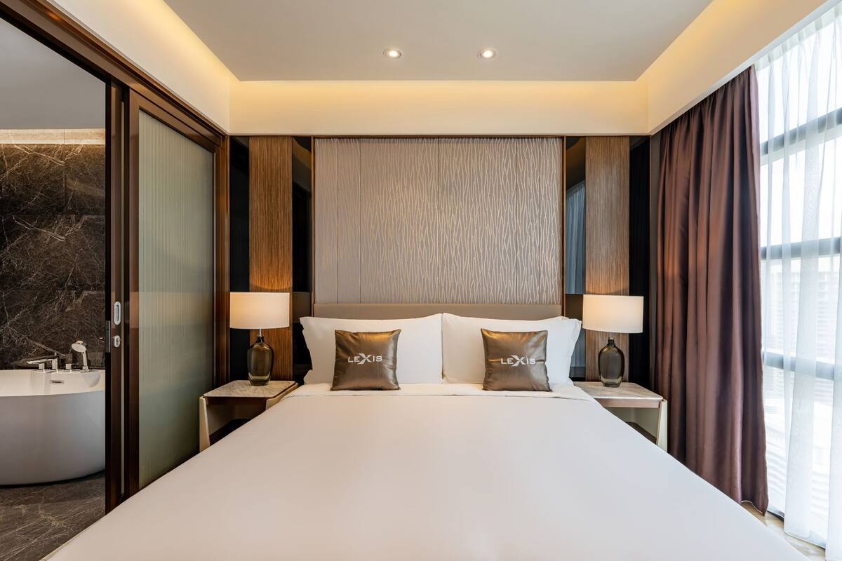 Een hotelkamer met een groot bed, twee bedlampjes, een hoofdeinde met een structuur en een uitzicht op de stad door het raam.