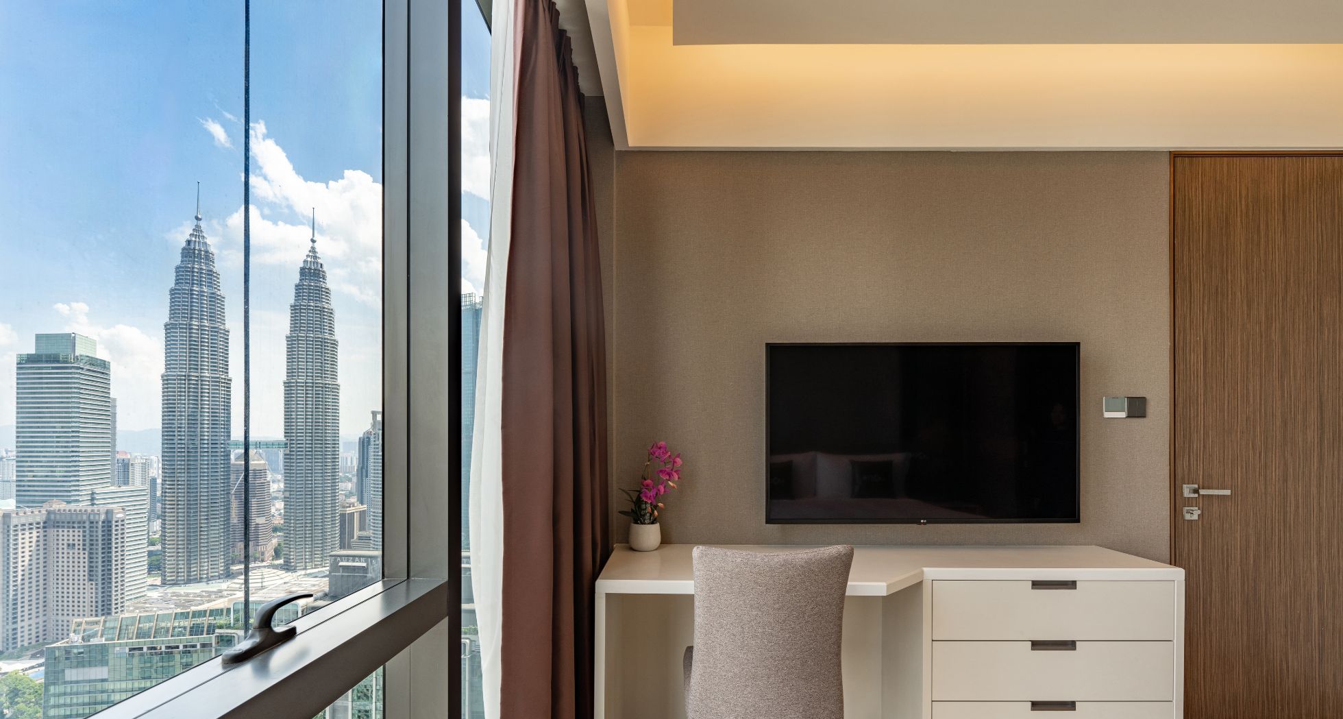 A modern hotel room with a large window, a flat-screen TV, a small desk, and a wardrobe.