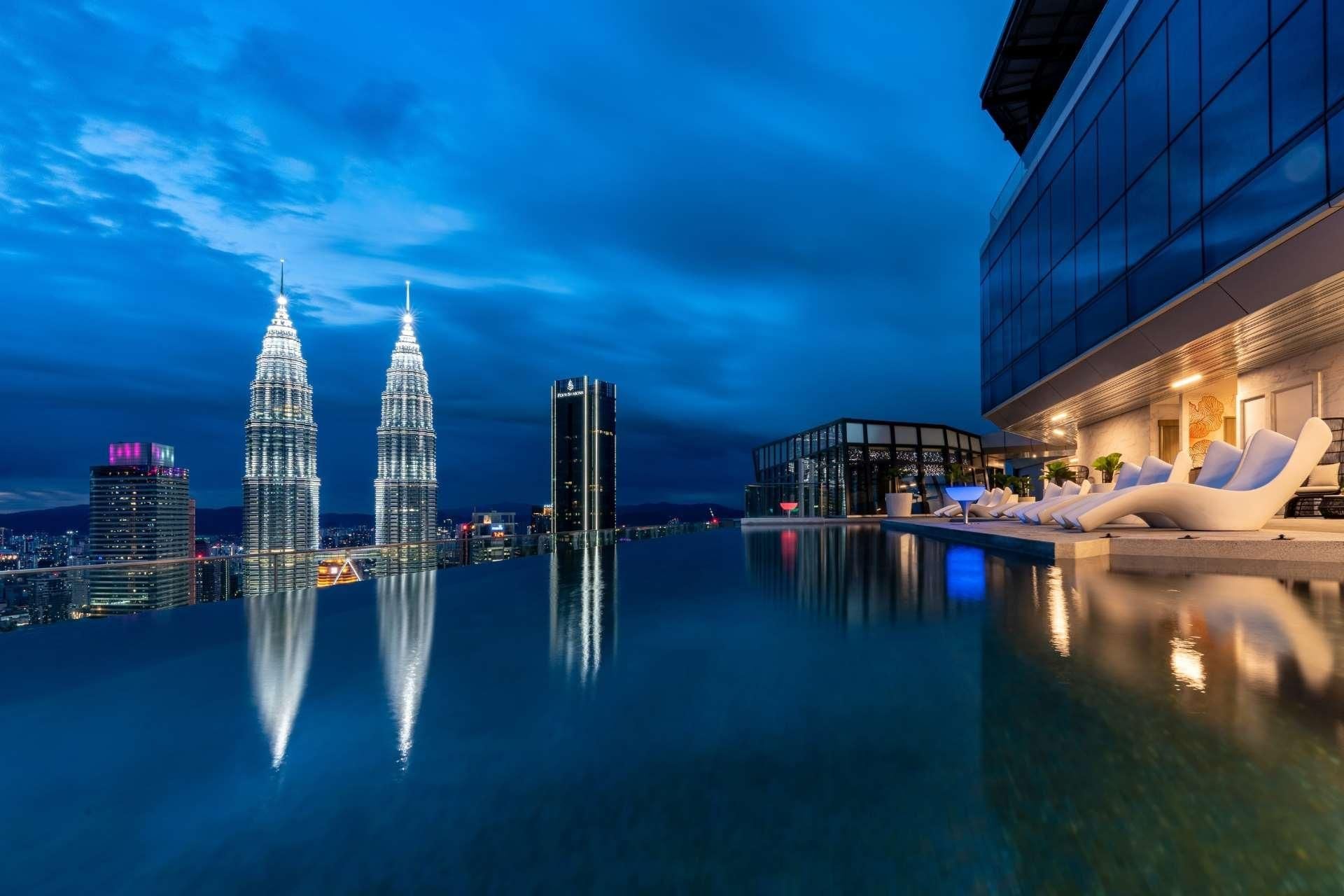 Een dakzwembad met oneindige horizon en uitzicht op de Petronas Towers, een modern gebouw met een glazen gevel, en een lounge met witte stoelen.