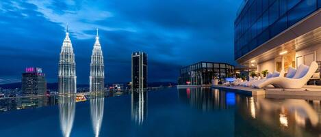 Een dakzwembad met oneindige horizon en uitzicht op de Petronas Towers, een modern gebouw met een glazen gevel, en een lounge met witte stoelen.