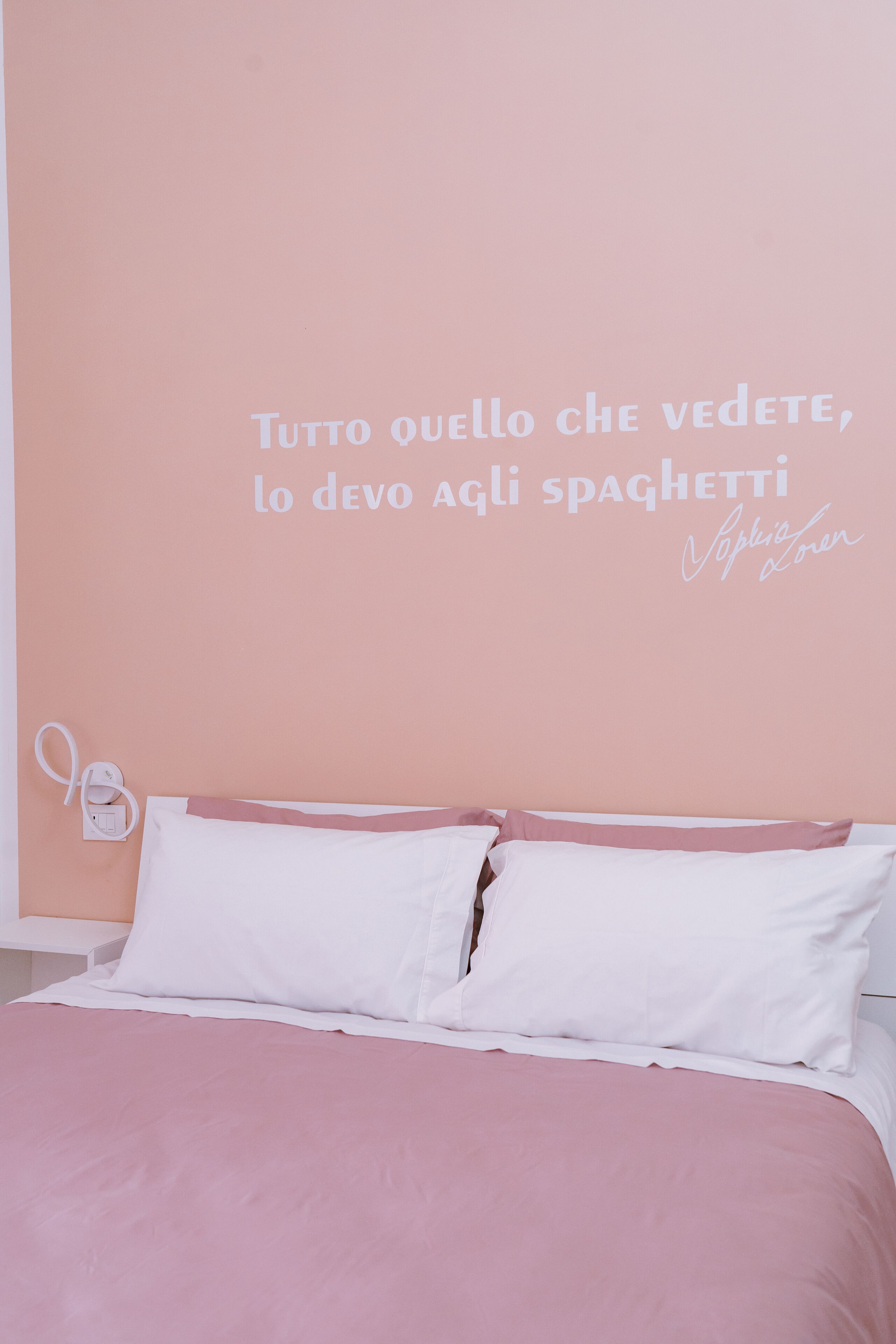 Un dormitorio con pared rosa, ropa de cama blanca y una cita en italiano.