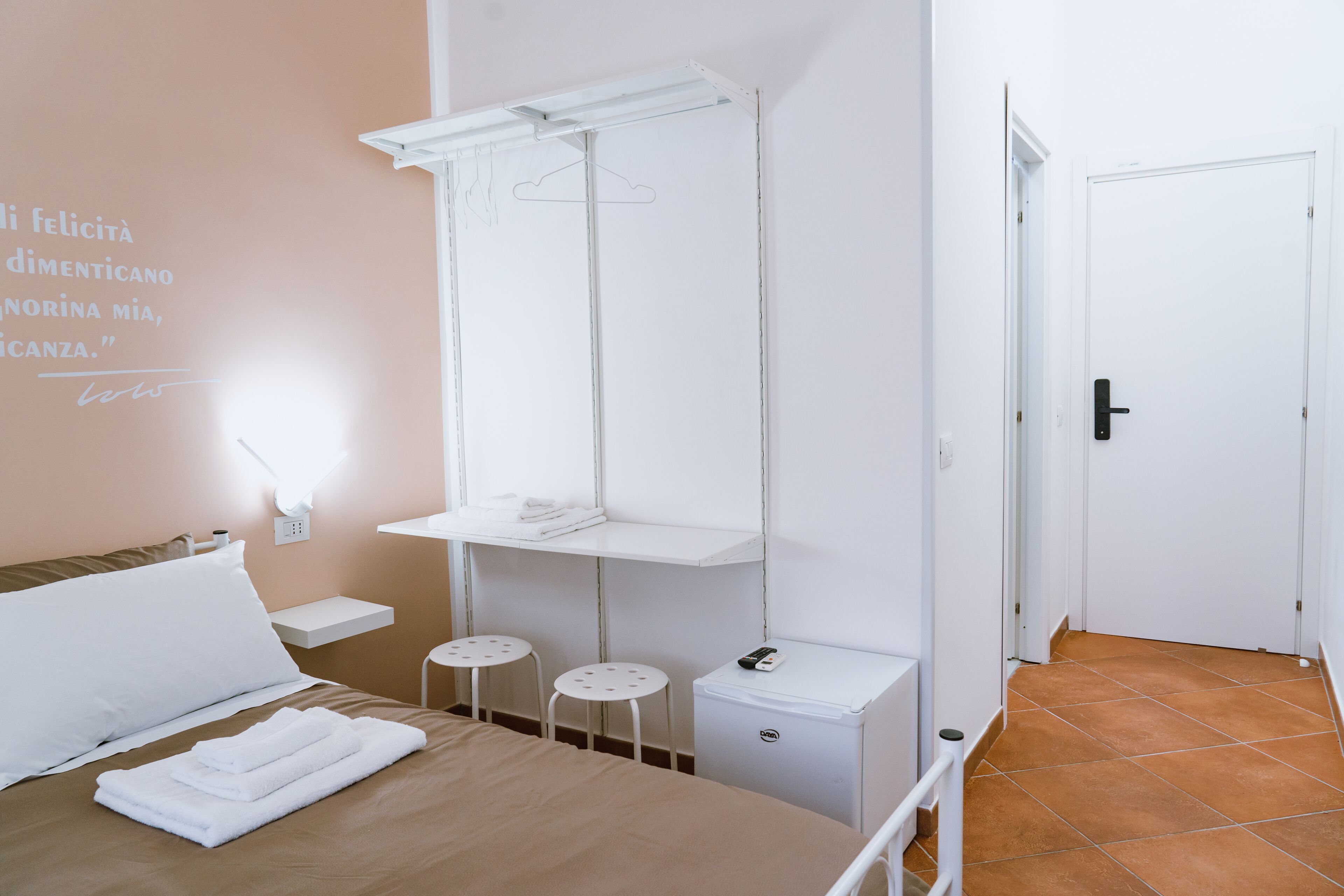 A hotel room with a bed, a bedside table, a small table, a chair, a wardrobe, a hanging light, and a door.