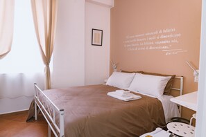 Een net opgemaakt bed met een bruine spreideken, een wit kussen en een opgevouwen handdoek ernaast. Op de muur hangt een lijst met een citaat en naast het bed staat een klein tafeltje met een stoel.