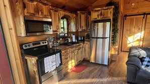 Interior de madera con cocina equipada, que incluye estufa, horno, microondas y refrigerador. Hay un ventilador de techo, armarios de madera y una alfombra roja en el suelo.