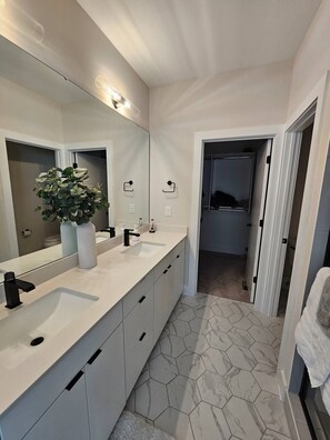 Een moderne badkamer met een grote spiegel, twee wastafels, een kamerplant en een vloer van honingraatmozaïek.