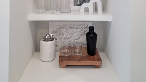 Una estantería blanca con una planta en maceta, vasos transparentes, un cuadro con una vista urbana y una bandeja de madera con una botella negra.