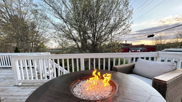 A round outdoor table with a built-in fire feature, surrounded by wicker furniture and a white deck railing.