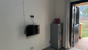 A room with a flat-screen TV mounted on the wall, a refrigerator with a sticker, and a door leading to an outdoor area.