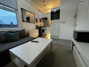 A compact living space with a sofa, a white coffee table, a kitchen area including a microwave, oven, and washing machine. - Onsite Apartments in Central Brentwood (Brentwood)