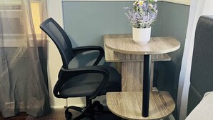 Um quarto pequeno com um sofá azul, uma cadeira de escritório preta, uma mesa lateral de madeira com uma planta em vaso e uma janela com persianas.