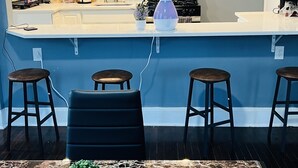 Uma cozinha moderna com armários brancos, micro-ondas de aço inoxidável e fogão a gás. Há dois banquetas de bar e uma pequena planta em vaso sobre o balcão.