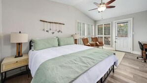 A bedroom with a bed, two chairs, a desk, a ceiling fan, and a window with shutters. - Y'all Stay Here Inn (Austin)