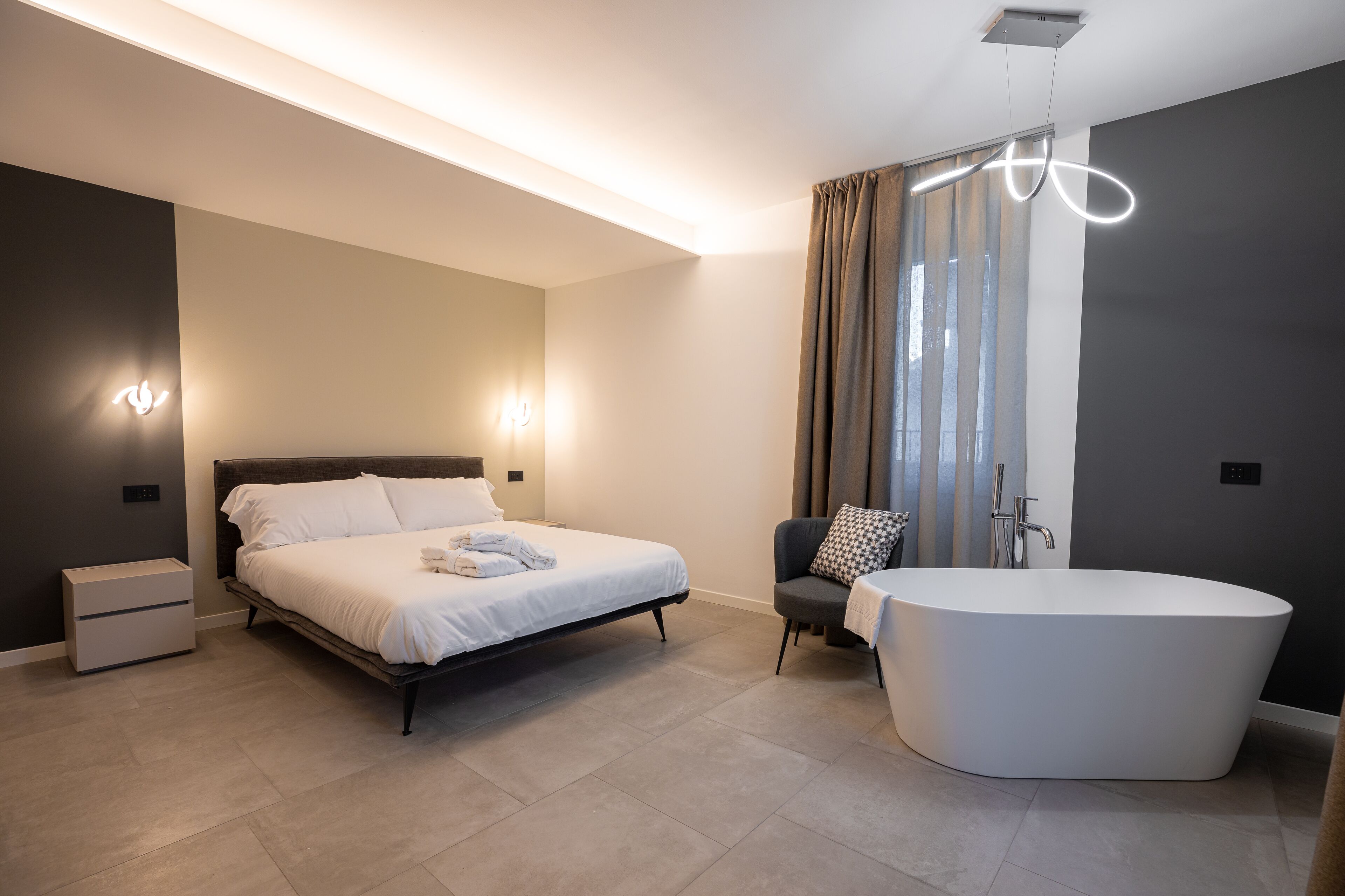 Ein modernes Schlafzimmer mit Bett, Nachttischen, einem Sessel, einer Badewanne und einer Deckenleuchte.