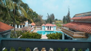 A pool area with a view of a building with a red-tiled roof, surrounded by greenery and palm trees. - Icon Heritage Bakkhali - Where the Sea Meets Luxury (Kakdwip)