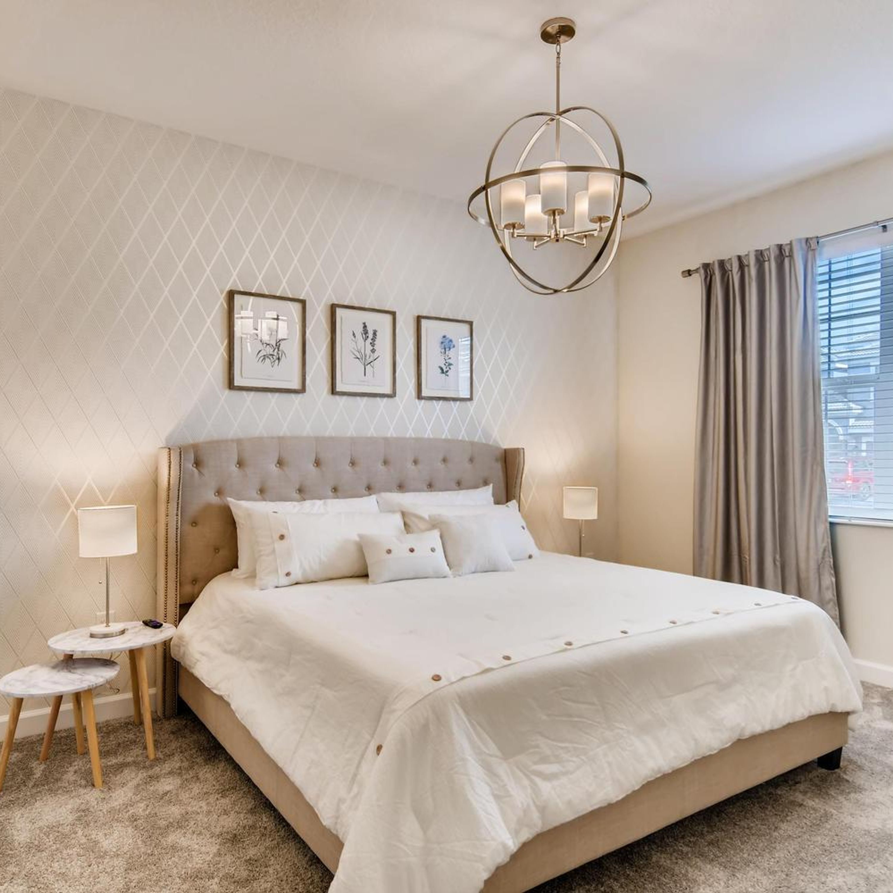 A spacious bedroom with a large bed, a tufted headboard, and a chandelier. There are three framed botanical prints on the wall, and a window with curtains.