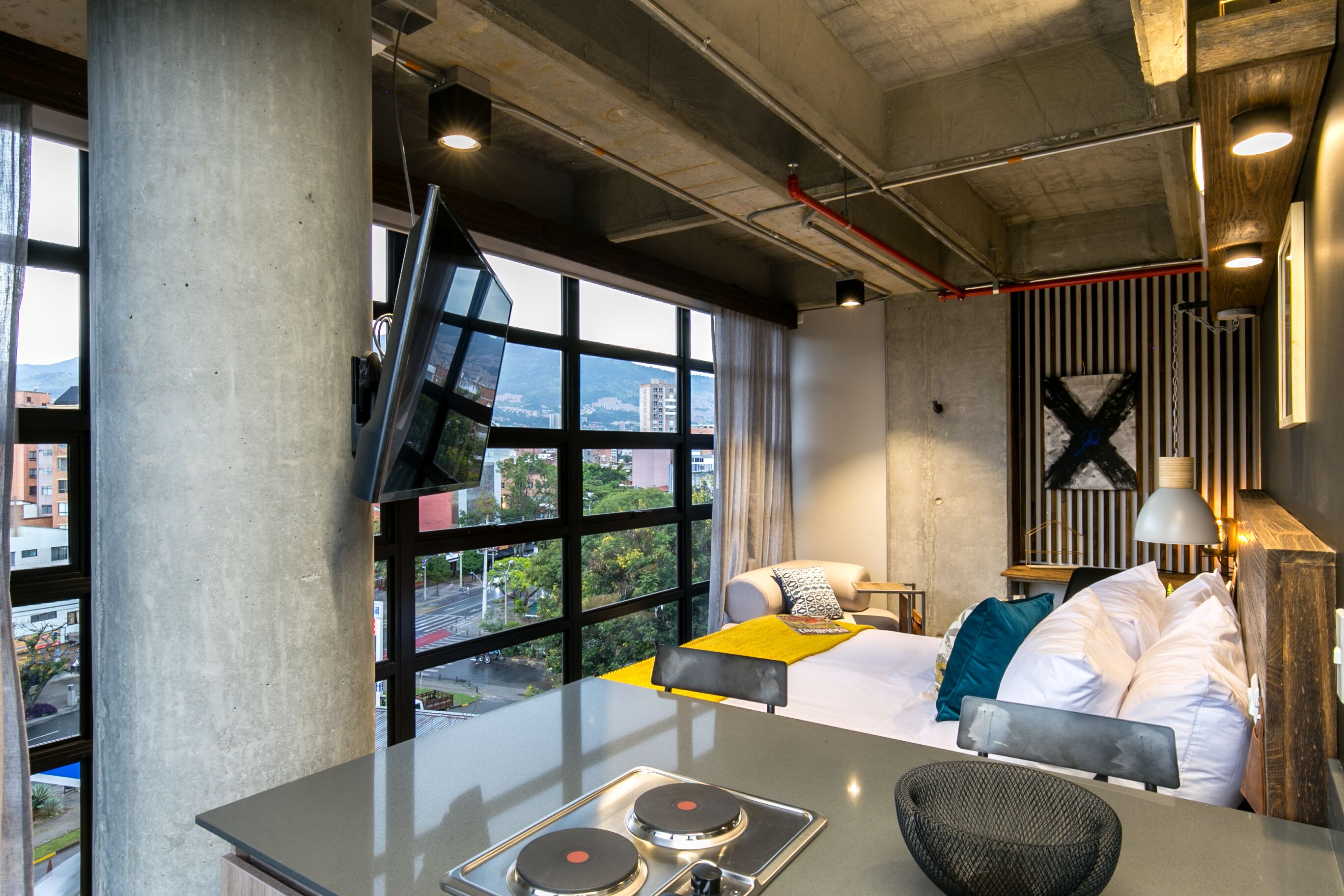 A modern hotel room with a bed, a small table, and a view of the city through large windows.
