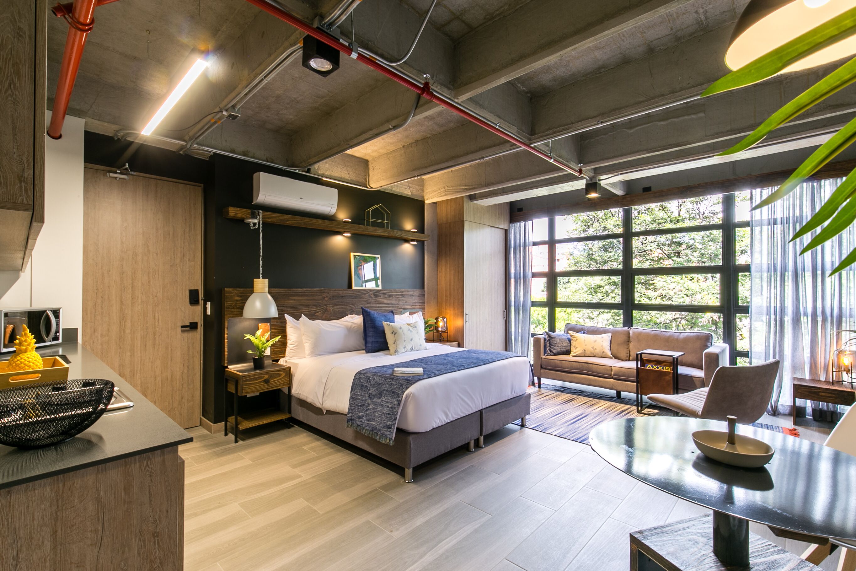 A modern hotel room with a large bed, a sitting area with a sofa and armchairs, a dining table, and a view of the outdoors through large windows.