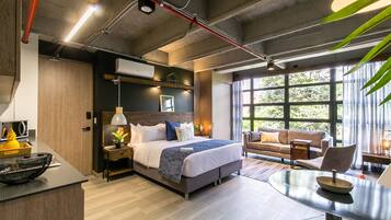 A modern hotel room with a large bed, a sitting area with a sofa and armchairs, a dining table, and a view of the outdoors through large windows.