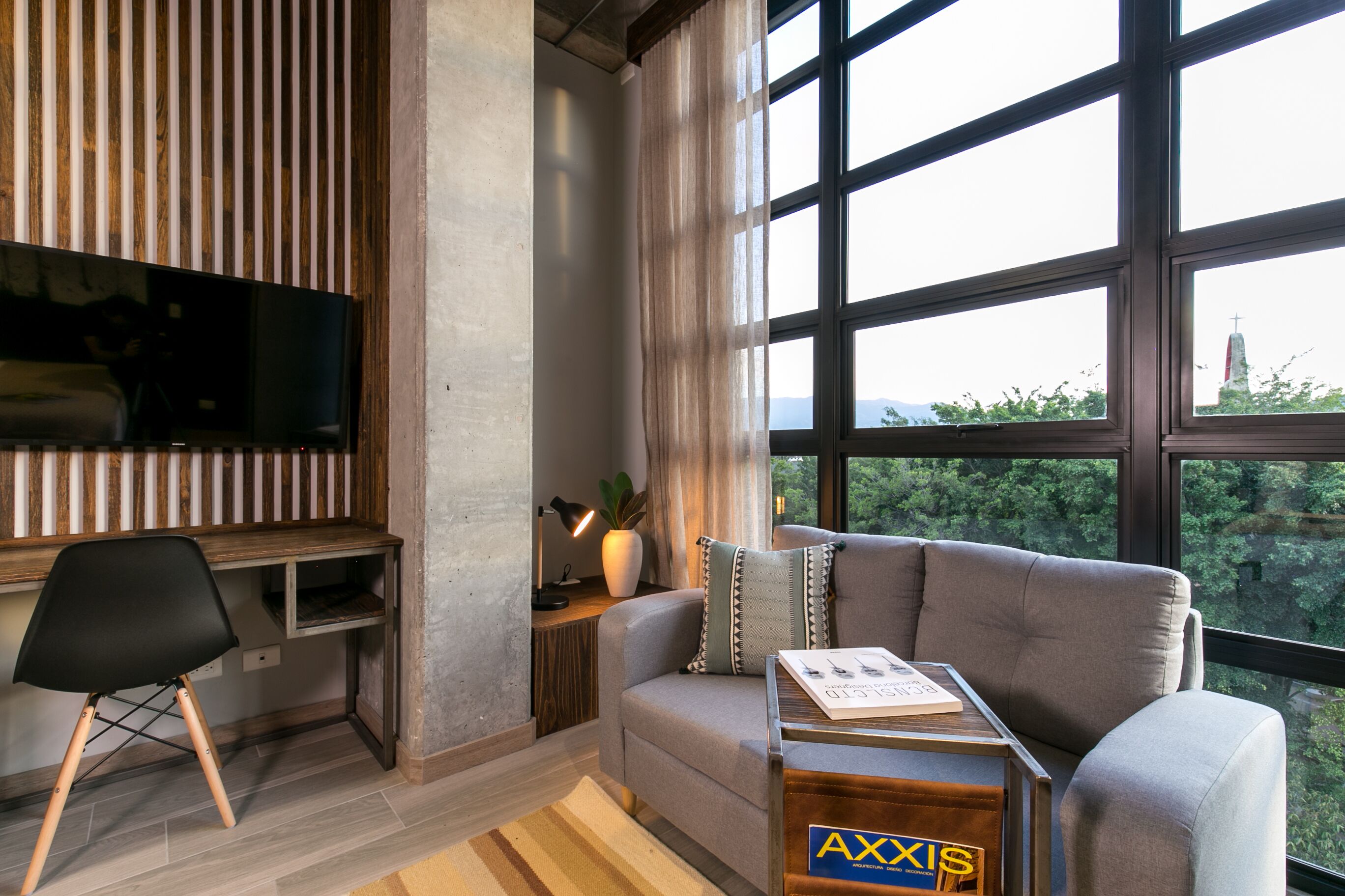 Una sala de estar moderna con un sofá gris, un escritorio de madera, una silla negra y un ventanal con vistas a follaje y una estructura distante.
